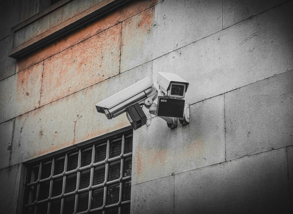 Two surveillance cameras mounted on an aged concrete wall beside a barred window, capturing urban security.